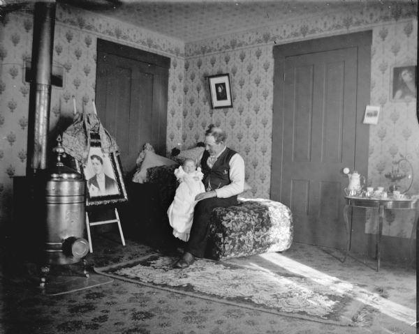 Portrait of a man posing sitting and holding an infant next to a woodstove. Next to the stove is a large portrait of a man on an easel.