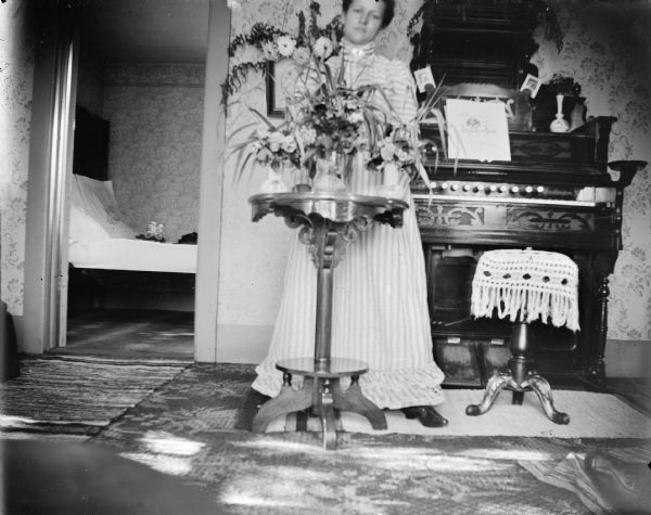 Portrait of a woman wearing a light-colored dress posing standing behind a plant stand with a flower arrangement. Behind her is an organ against the wall. Through a doorway on the left is a bed.