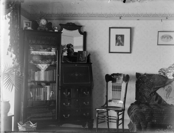 View of a bedroom, with a bookshelf, dresser, wooden chair, bed, and several framed photographs. There is a plant in front of a window on the far left.