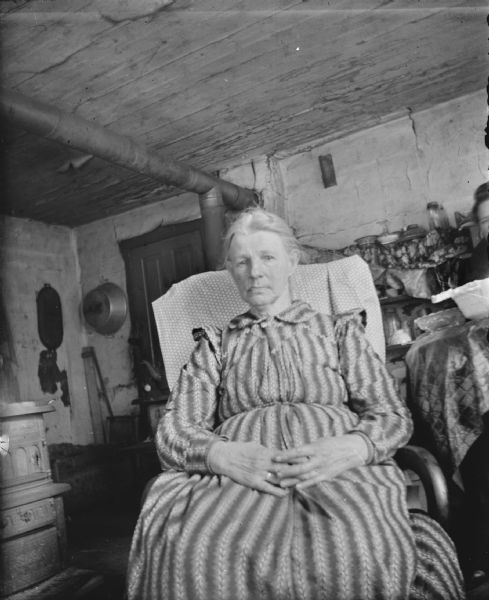 Portrait of a woman wearing a striped dress posing sitting in a chair near a wood stove. Stove piping is above her in the background, and dishes are on tables and shelves behind her on the right.