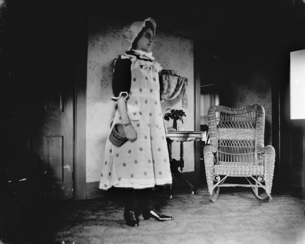 Portrait of a woman wearing a light-colored polka-dot dress and bonnet posing standing in a room. Behind her is a wicker rocking chair and a small table.