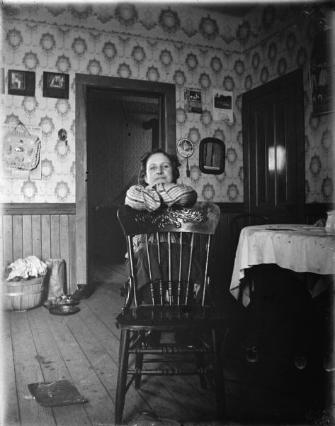 A woman is posing sitting on a chair, with her arms and feet on the back of a wooden chair in front of her. On the right is a table. The room has wallpaper on the walls, and two doorways.