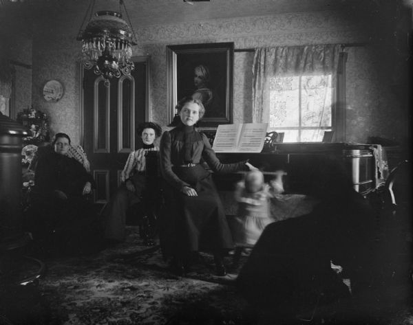 Group portrait of three women posing sitting in a parlor. One of the women is sitting in front of a piano. Next to her is a small child (blurred by movement) reaching up to the keys on the piano. In the foreground on the left is a wood burning stove.