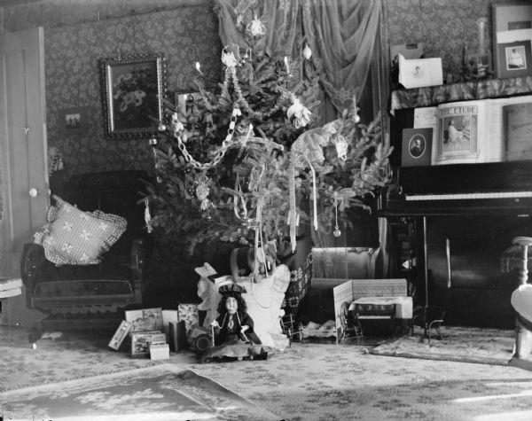 View of a living room with a decorated Christmas tree in between a piano and an upholstered chair. Toys are arranged beneath the tree.