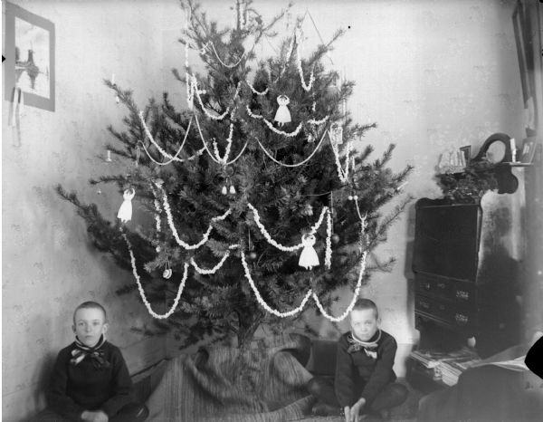Portrait of two boys posing sitting underneath a decorated Christmas tree set up in the corner of a room next to a wooden dresser. The tree is decorated with strings of popcorn, and angels.
