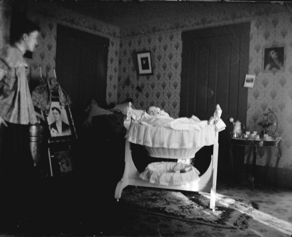 View of a woman standing on the left next to a wood burning stove. She is looking at an infant in a bassinet in the center of a room. A large portrait of a man is on an easel just behind the stove.