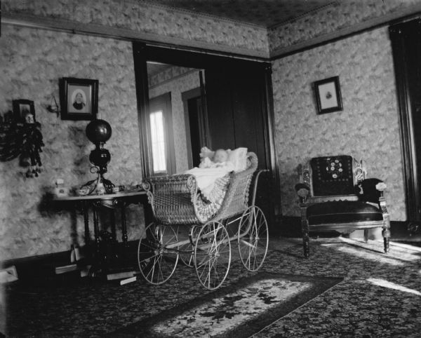 An infant is sitting in a baby carriage in a room with wallpapered walls. On the right is an upholstered chair, and on the left is a table with a large lamp. Two framed portraits are hanging on the walls.
