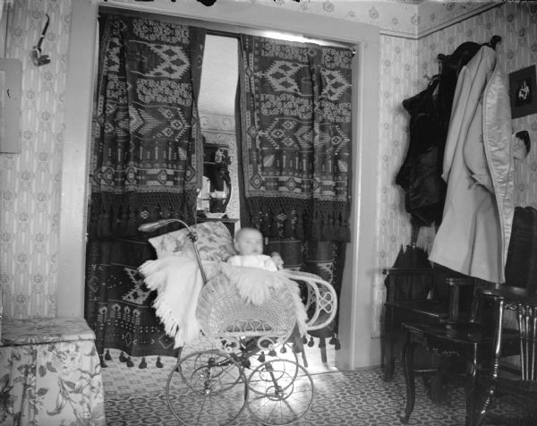 An infant is sitting in a baby carriage in a room that has coats and umbrellas hanging near a chair on the right. Behind the infant is a curtain hanging in a doorway.