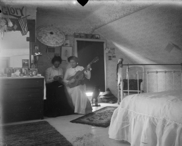 View across bedroom towards two women sitting together. The woman on the right is holding a guitar. On the left is a dresser with a mirror, and a bed in the right foreground. A stuffed owl on a stand is on the floor in front of a window with a dark shade.