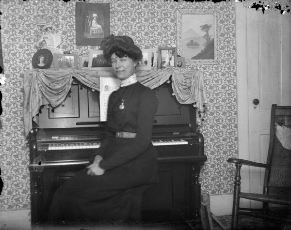 Portrait of a woman posing sitting on a bench in front of a piano. On top of the piano are numerous framed photographs, and two prints are hanging on the wallpapered wall.