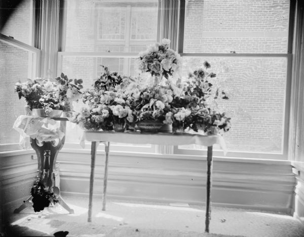 Flower arrangements on display on top of two tables in front of large windows in the corner of a room. A brick building is visible through the windows. Identified as the interior of the Richards House, with the Methodist Church visible through the windows. The flowers are probably for the graduation of Edna Richards Turner from high school in 1897.