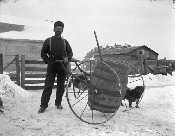 Outdoor portrait of a man posing standing. He is holding a cart made of a barrel with two large metal wheels. Identified as probably a pig swill cart. A dog is standing just behind the barrel on the snow-covered ground. There is a fence, barn and other wooden buildings in the background.