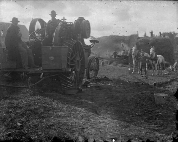 In the foreground on the left are two men sitting on machinery belt driving the thresher. In the background, a man is standing and holding the bridles of two horses. Other men are working in the far background around the threshing machine set up near a large pile of hay.