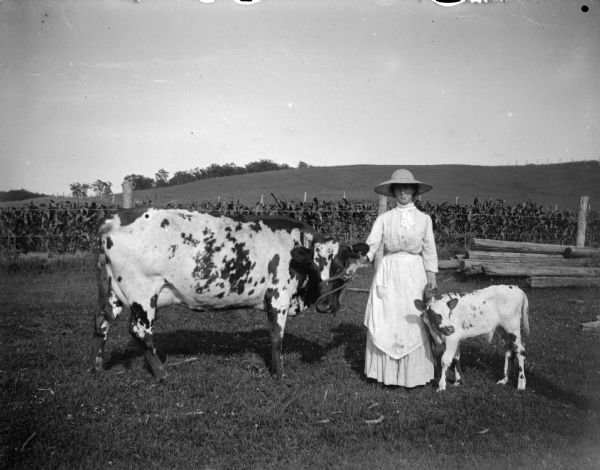 Outdoor portrait of a woman wearing a light-colored dress, apron, and a hat. She is standing holding a cow and a calf. In the background is a cornfield and a hill.