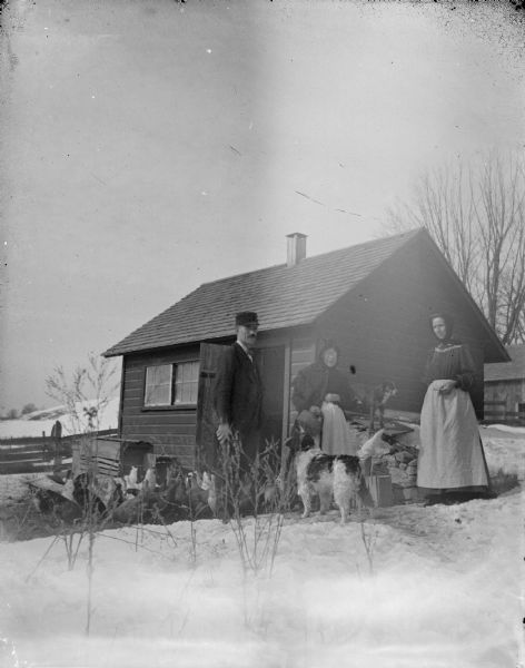 Group portrait of a man, two women, and two dogs posing standing among chickens on the snow-covered ground in front of a small wooden building, perhaps a chicken coop.
