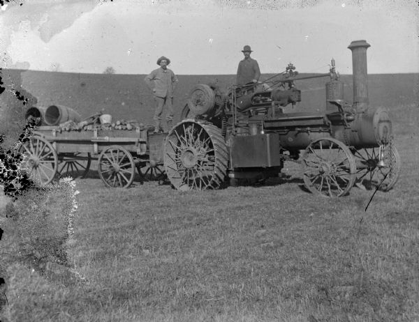 View of two men posing standing on a tractor and wagon in a field.