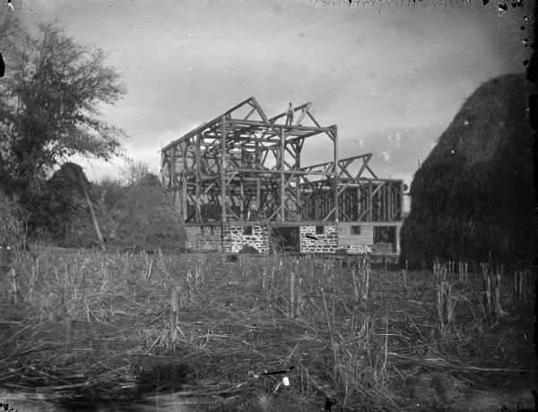 View across field towards two men posing standing on the frame of a barn under construction next to a large haystack.