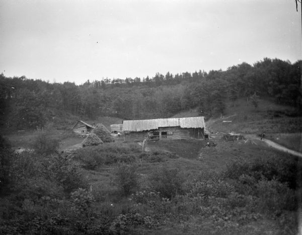 Elevated view down hill towards a wooden building in the distance. There are haystacks next to the building. There is a forested hill in the background.