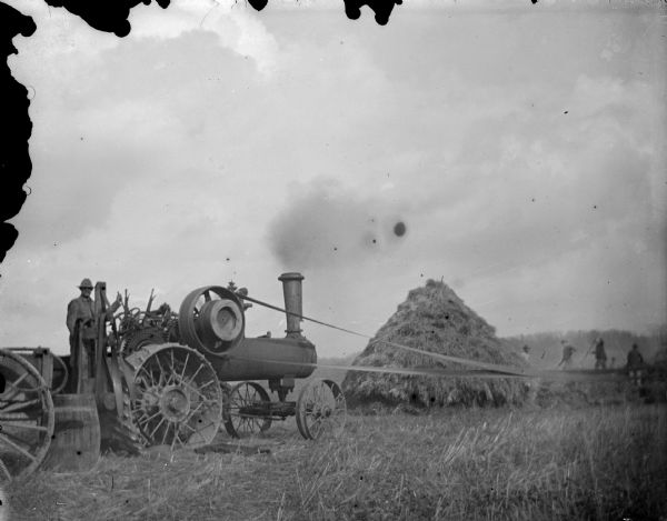 Exterior portrait of a man posed standing on a tractor running threshing machinery in the distance next to a group of men and a haystack.