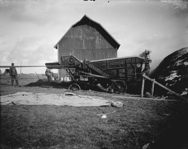 View across barnyard towards men standing next to threshing machinery marked "Aultman-Taylor" in front of a wooden barn.
