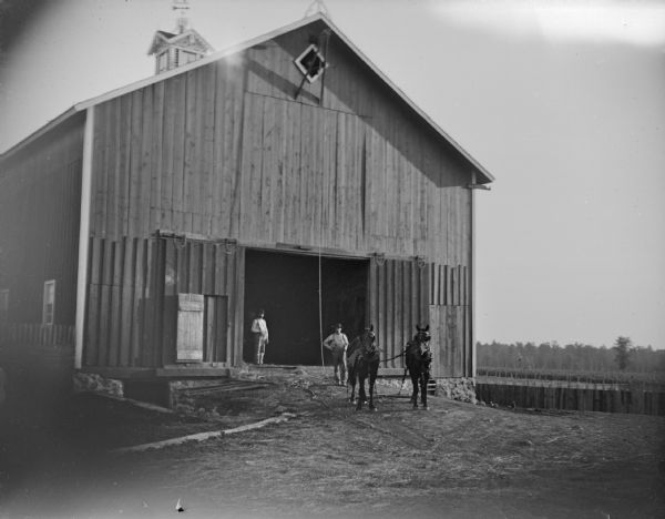 View across barnyard of two men posing standing near a team of two horses in front of a large wooden barn.