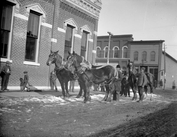View down street towards a man displaying a team of two horses, a man displaying a single horse, and boys standing on the sidewalk in front of a large brick building. Location identified as the intersection of Main and First Streets.