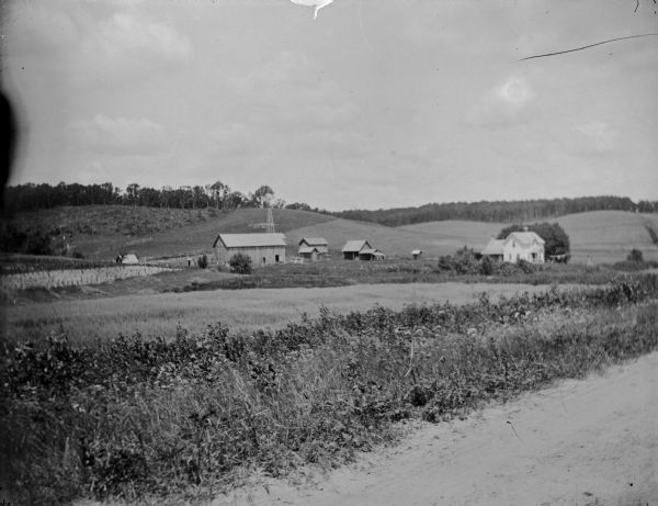 Exterior portrait of wooden buildings and a windmill in the distance in front of rolling countryside taken from a road.