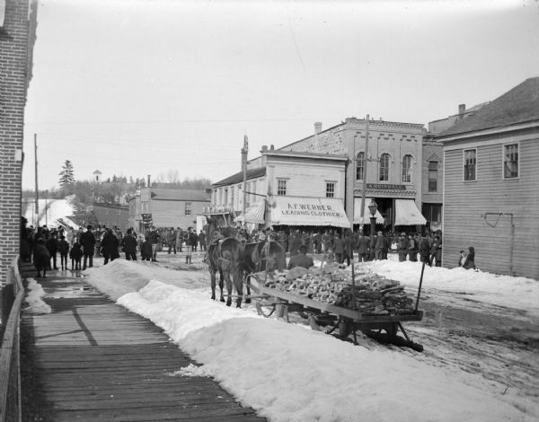 View down sidewalk towards people lining a snow-covered city street. There is a sled loaded with wood in the foreground. Location identified as looking north toward the corner of Main and First Streets.