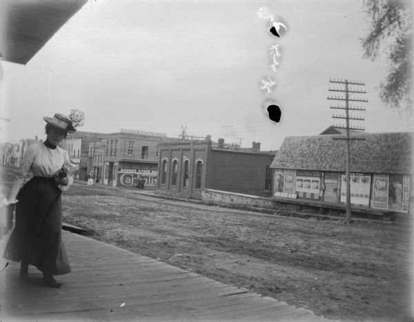 View down board sidewalk towards a woman wearing a light-colored blouse and dark-colored skirt walking on the left along an unpaved city street. Location identified as Main Street, looking toward First Street. There are signs and posters on the side of a building across the street on the left. Some of the signs read: "John Deere Plows," and "Quo Vadis."