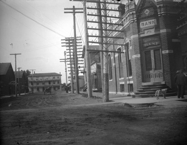 View down unpaved street towards an intersection of two city streets. The Jackson County Bank is located on the right, and utility poles line the right side of the street. Location identified as the intersection of Main and First Streets in Black River Falls looking south down First Street.