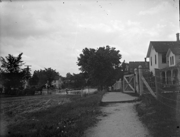 View down unpaved street towards flooded area in the distance. On the street is a girl walking towards the water, and on the left is a group of people near horse-drawn wagons. Location identified as South Second Street between Pierce and Lincoln Streets on the West side. On the right further down the board sidewalk is another group of people looking at the flood.