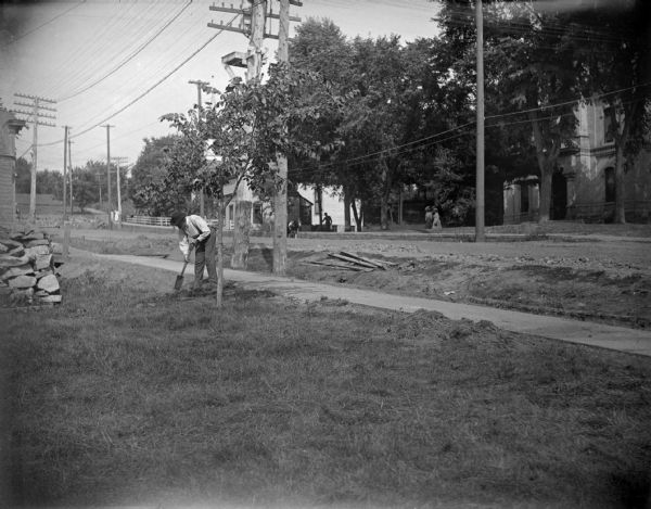 View across lawn towards a man working with a small spade on the left, and men and women in the distance across a city street. Location identified as Main Street in front of the Jackson County Court House in Black River Falls, which is visible on the right.