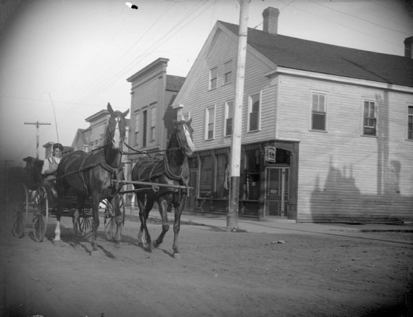View from street towards a team of horses being driven by a woman sitting in a carriage on a city street. Location identified as the southeast corner at the intersection of Main and First Streets. Storefronts are in the background along the sidewalk.