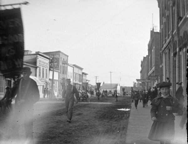 View from sidewalk looking across unpaved street, with men, women, and children following along with a procession. A young man in the left foreground is holding a banner. Location identified as Water Street looking south.