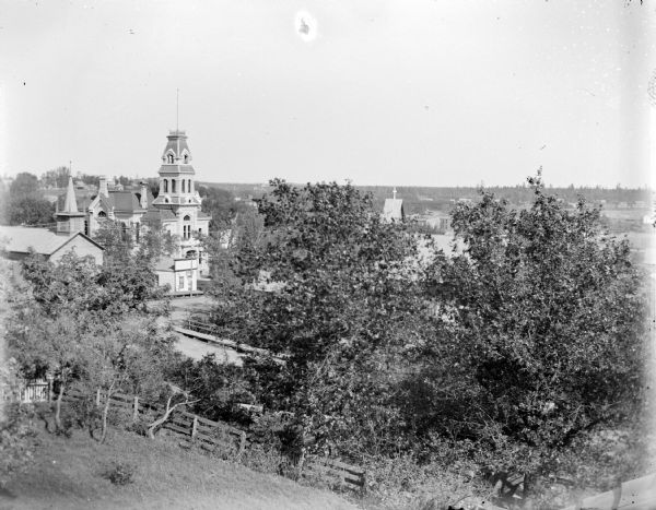 Elevated view of town, with trees and a wooden fence around a yard in the foreground. Location identified as Main Street, the Baptist Church is on the left and the Jackson County Courthouse is left center.