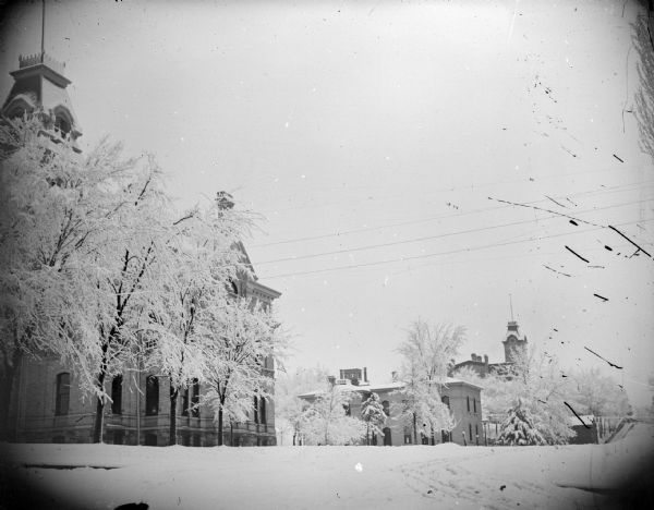 View towards trees and buildings lining a snow-covered city street. Location identified as Third Street in Black River Falls, the Jackson County Courthouse on the left, the County jail center, and the school on the right.