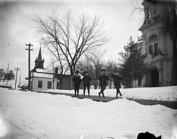 View from street towards four boys posing standing on a wooden sidewalk lining a snow-covered street. Location identified as the Jackson County Courthouse on the right, the office of Dr. Gebhardt in the center, and the Baptist Church in the distance.