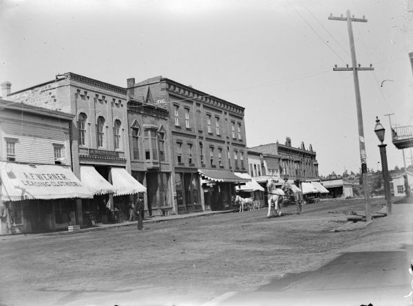 View across street towards a man sitting in a buggy pulled by a single horse coming down a city street. There are storefronts along the opposite side of the street along the sidewalk. A man is standing in the unpaved street near a storefront with an awning that reads: "A.F. Werner, Leading Clothier." Location identified as the intersection of Main and First Streets looking east.