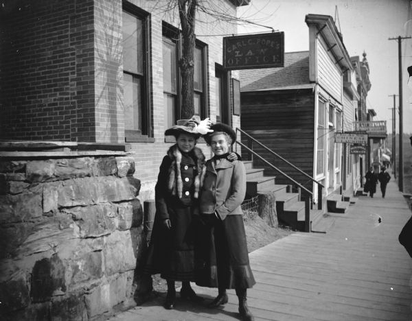 View down wood sidewalk towards two women posing standing in front of the Law Office of Carl C. Pope, with other storefronts in the background. Location identified as the north side of Main Street between Second and Third Streets.