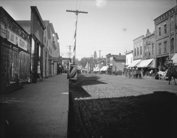 View down sidewalk. On the left is a wall with signs and posters, and further down are storefronts, including the Lockens Shoe Store. On the other side of the street is a procession of people along the sidewalks along storefronts, some with awnings. Identified as Main Street looking west from Water Street.