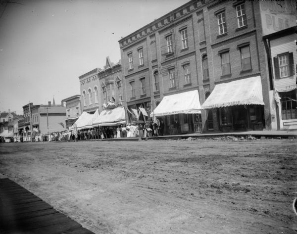 View across unpaved street towards a large group of men and women walking down a wooden sidewalk along a city street. Location identified as Main Street. Flags are displayed on signs, buildings, and storefronts. The storefronts, some with large awnings, are between Water and First Streets on the north side of the street.