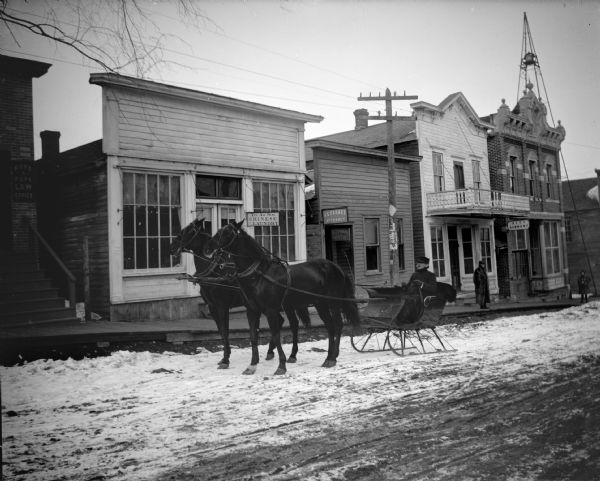 View across street towards a man posing sitting in a sleigh pulled by a team of two horses on a snow-covered city street. Location identified as the north side of Main Street in Black River Falls, in front of the Yep Ah Sing Chinese Laundry. On the left is a building a sign for the Pope & Pope Law Office, and on the right are signs for J.C. Forbes Attorney, and one for Dentist. Further down the street on the right is a sign for the City Library. Behind the brick building on the corner is a tall tower with a bell.