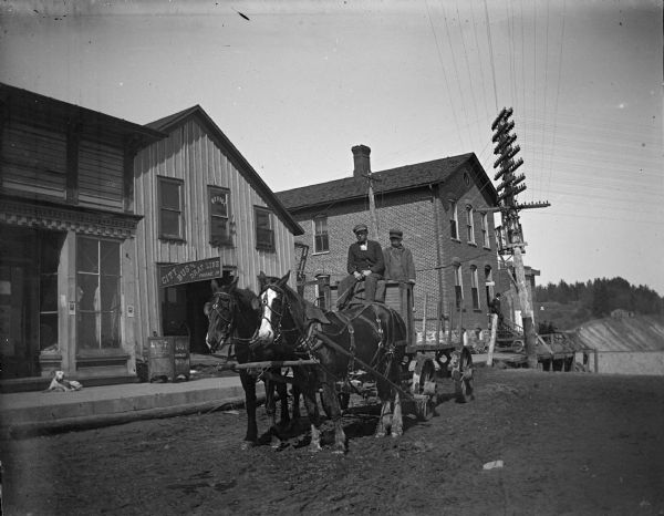 View down unpaved street towards two men posing sitting high in a wagon with a team of two horses in front of them. Location identified as South First Street in front of the bus line office and the telephone exchange building.