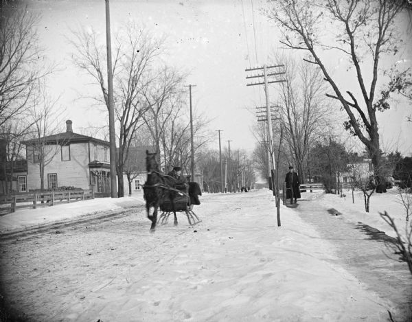 View down snowy street towards a horse pulling a man in a sleigh. There is a man walking down the sidewalk on the right. Location identified as the intersection of East and Eighth Streets, looking east, with the house of the McGillivray family on the left.