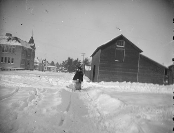 View down snow-covered path towards a woman pushing a child in a sleigh buggy. Location identified as probably looking from Third Street past the 1897 school to Jackson Street.
