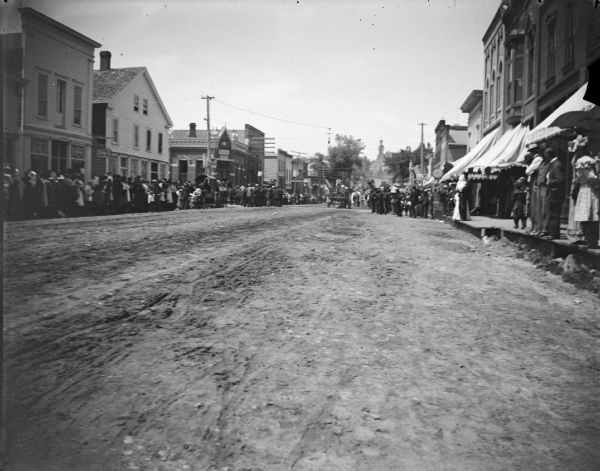 View down unpaved street towards large groups of people gathered on the sidewalks. Location identified as Main Street looking west from Water Street.