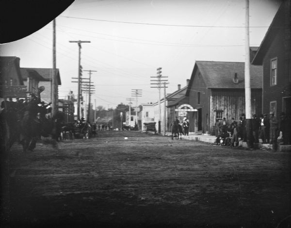 View down unpaved city street towards a man on the far left riding a horse. People are watching from wagons and along the wooden sidewalks on both sides of the street in front of storefronts. The City Livery building is further down the street on the right. Location identified as South First Street looking north from Fillmore Street.