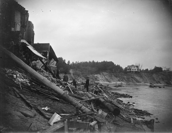 View along steep shoreline towards people among the rubble near buildings at the top of the bank on the left. The river is on the right. Location identified as the rubble remaining after a flood of the Black River washed away land up to South First Street in October 1922. In the distance on the right is the house of the Nelson family which had an undermined foundation and washed down the river one week after the flood.