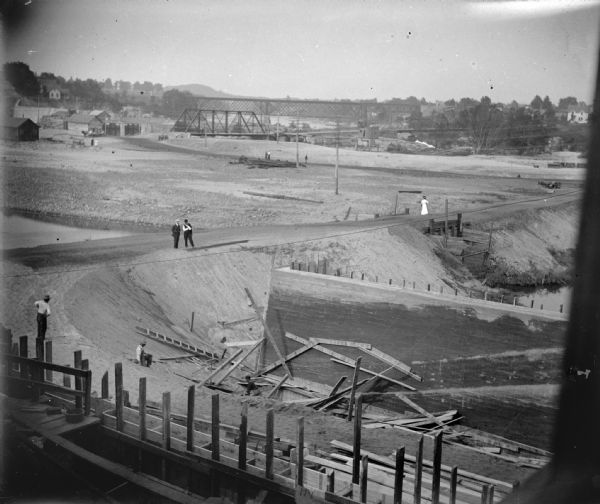 Elevated view of a construction site. There are two men standing on a dirt road, looking down at other men down below them working around a brick wall. A woman is walking along the road in the distance. Location identified as the fill and reconstruction after the 1911 flood in Black River Falls.