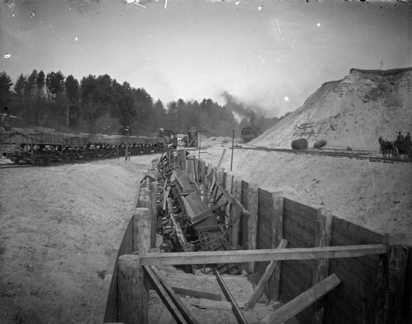 View of a construction site, with a ditch in the foreground. Location identified as the fill and reconstruction of Town Creek after the 1911 flood in Black River Falls. On the far right are two people in a horse-drawn wagon along railroad tracks, and in the distance a steam shovel billowing smoke while working just behind the base of a hill. On the left is a smaller locomotive with a string of railroad cars behind it.